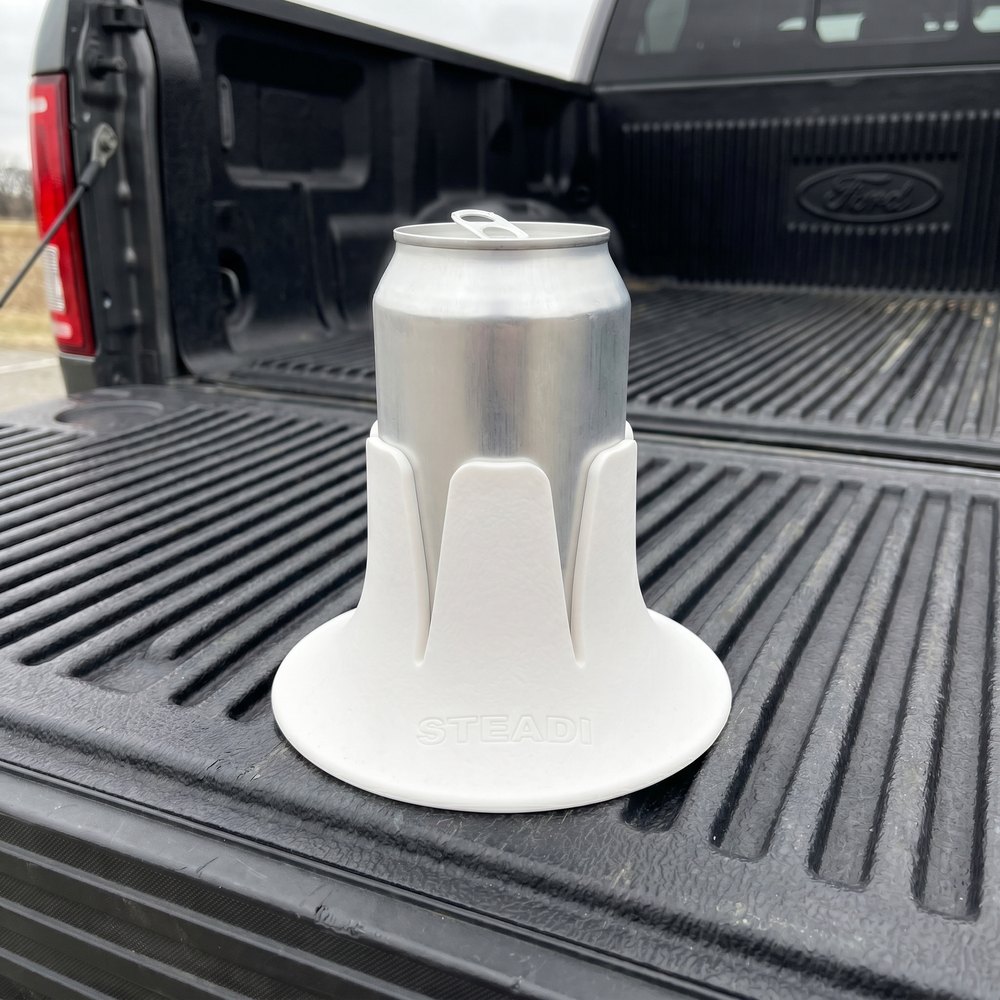 A white Steadi holding a can of beer, standing on the ribbed black bed rail of a pickup with the tailgate down behind it.