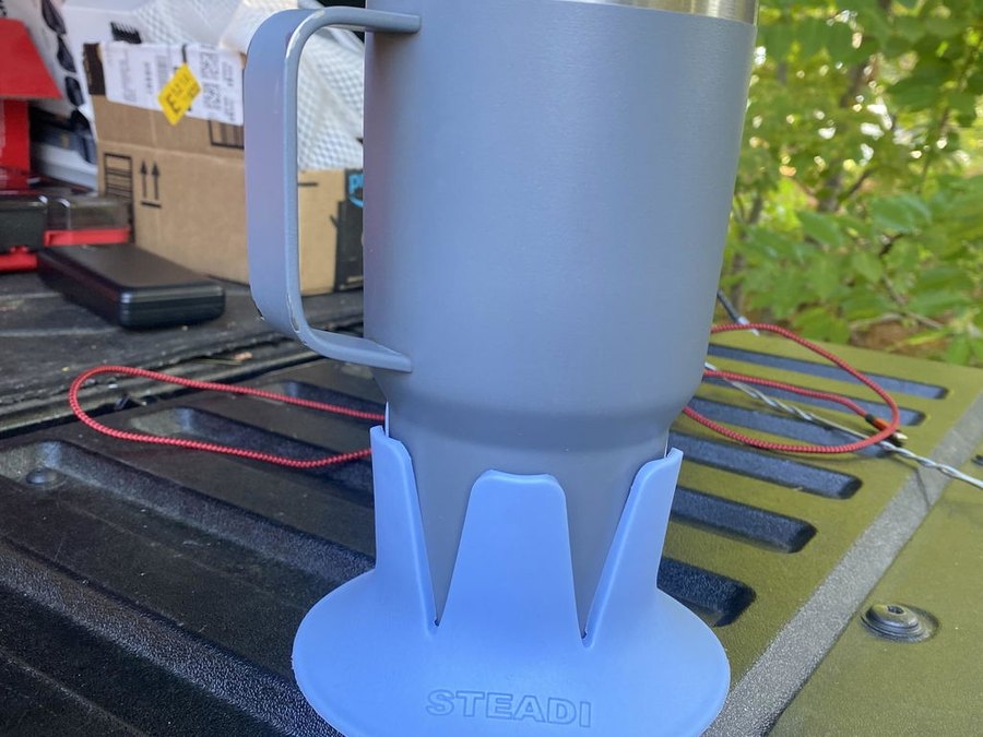 A grey insulated tumbler with a handle in a periwinkle STEADI base standing on the black plastic bed rail of a pickup truck, cargo and wiring in the bed behind, green trees beyond